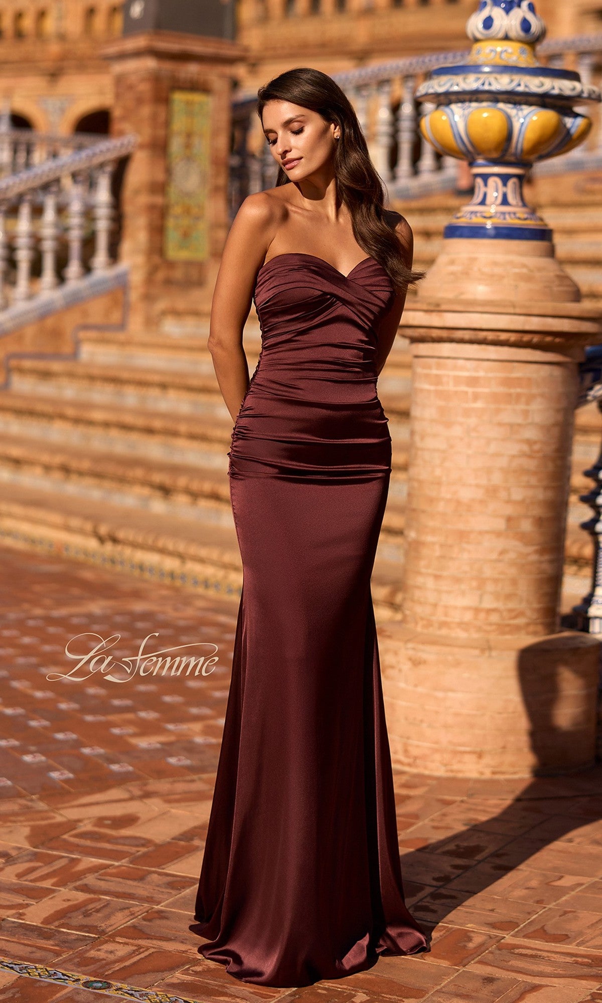 A woman in the chocolate La Femme 33531 classic strapless long prom dress with a ruched bodice stands outdoors on patterned brick steps, ornate railings, and decorative stonework in the background.